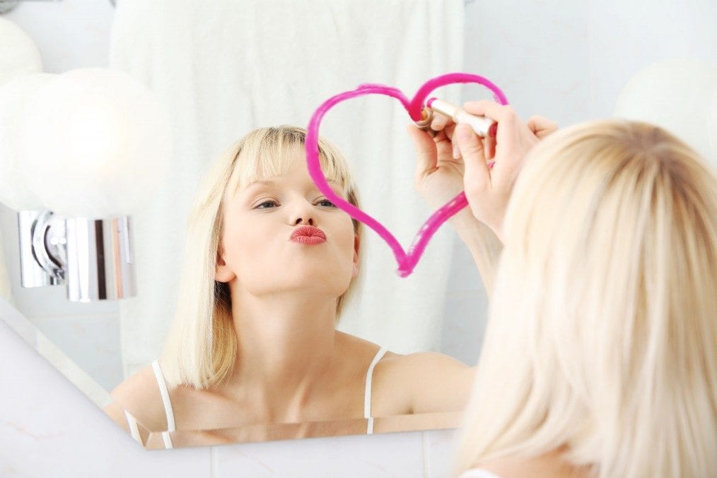 blonde woman drawing love heart in mirror with lipstick