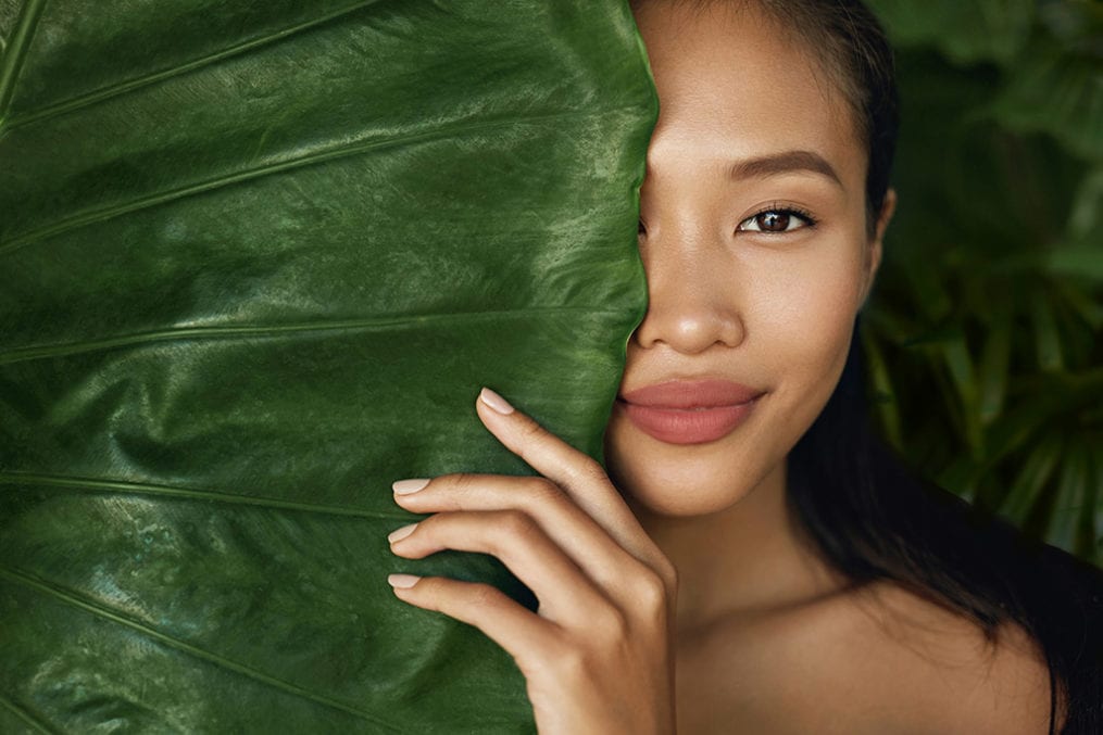 Woman model with natural makeup and healthy skin behind green leaf plant